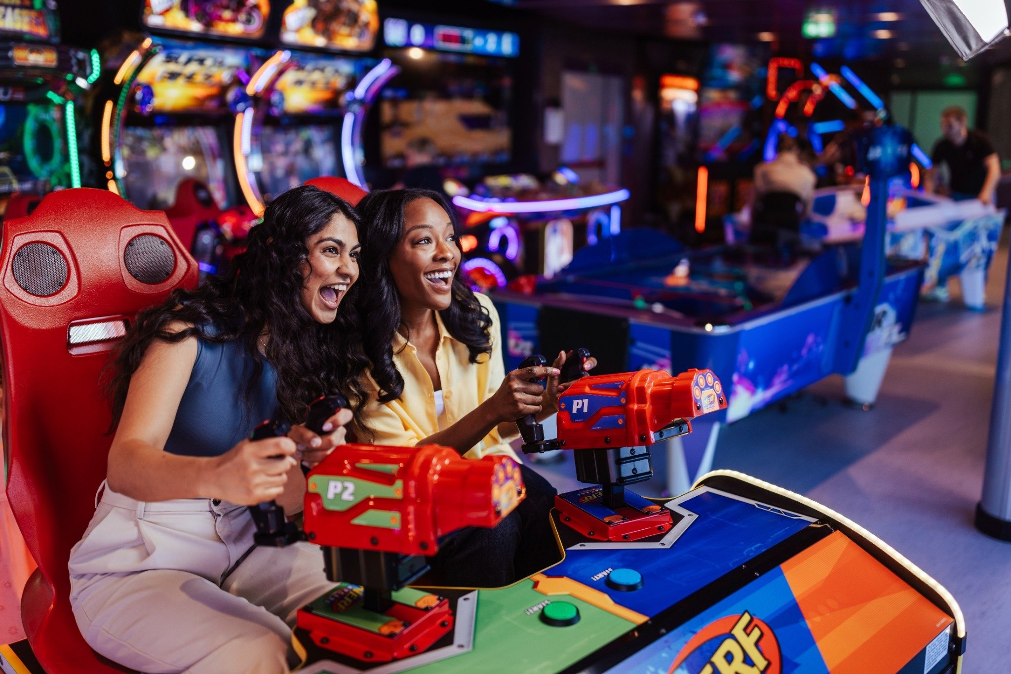 Duas mulheres se divertem jogando em máquinas de arcade coloridas, sorrindo animadas. | MSC Cruzeiros Duas mulheres se divertem jogando em máquinas de arcade coloridas, sorrindo animadas. | MSC Cruzeiros