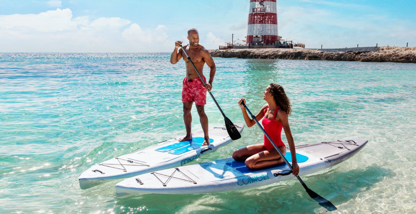 Two people paddleboarding in turquoise water near a red-and-white lighthouse | MSC Cruises Two people paddleboarding in turquoise water near a red-and-white lighthouse | MSC Cruises